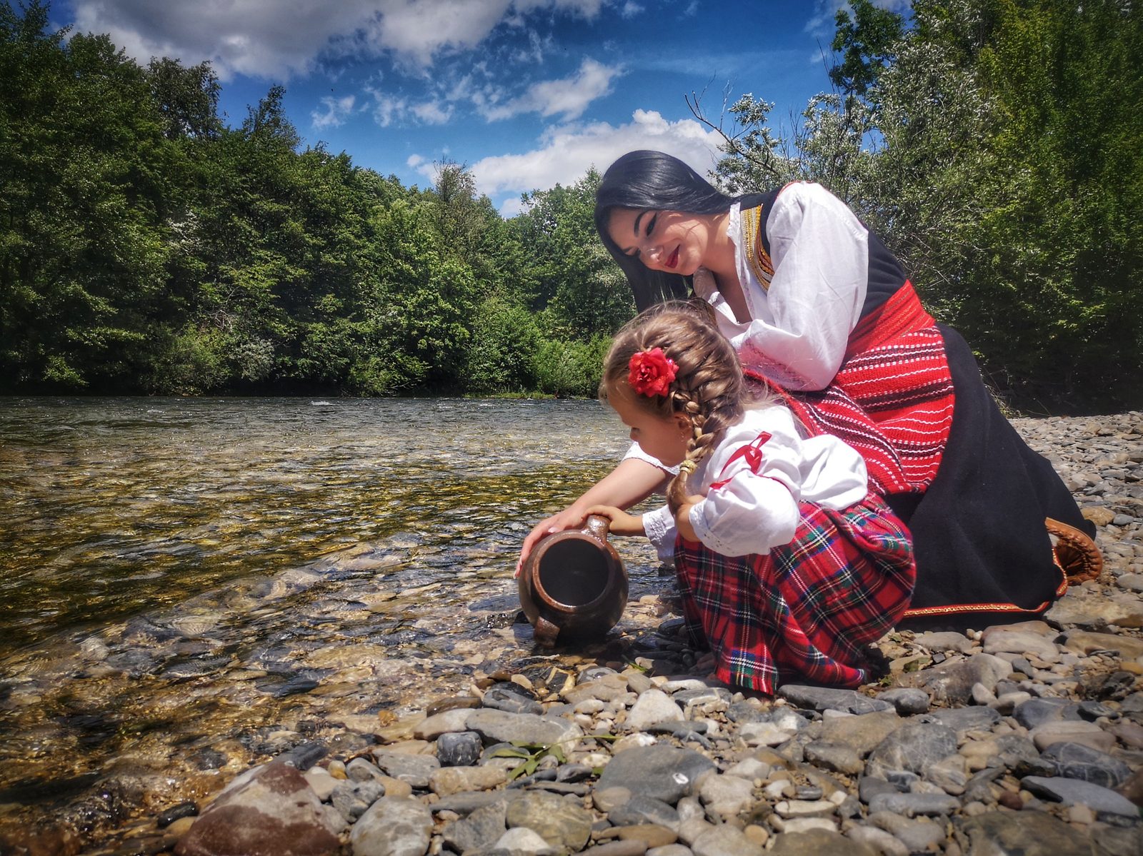 A woman and a young girl in traditional dress draw water from a clear river