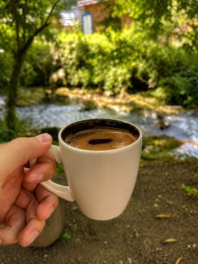 A cup of coffee in hand, with sun and the Janj stream in the background